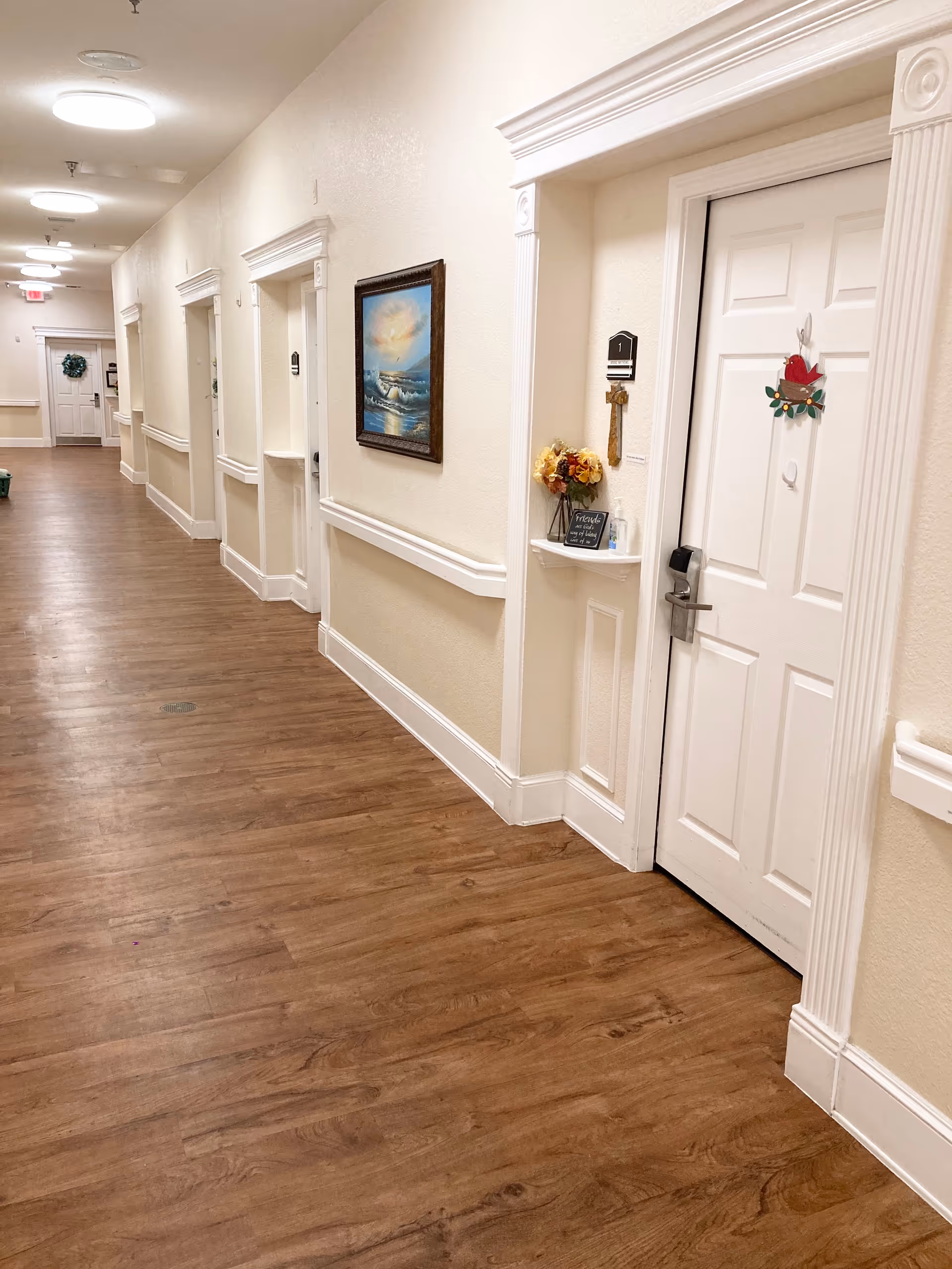 A long, well-lit hallway in an assisted living facility with wood-patterned flooring and cream-colored walls. Several white doors line the hallway, each with decorative trim and some with small decorations. A framed painting of a seascape hangs on the wall. The hallway has ceiling lights and a handrail running along the wall.