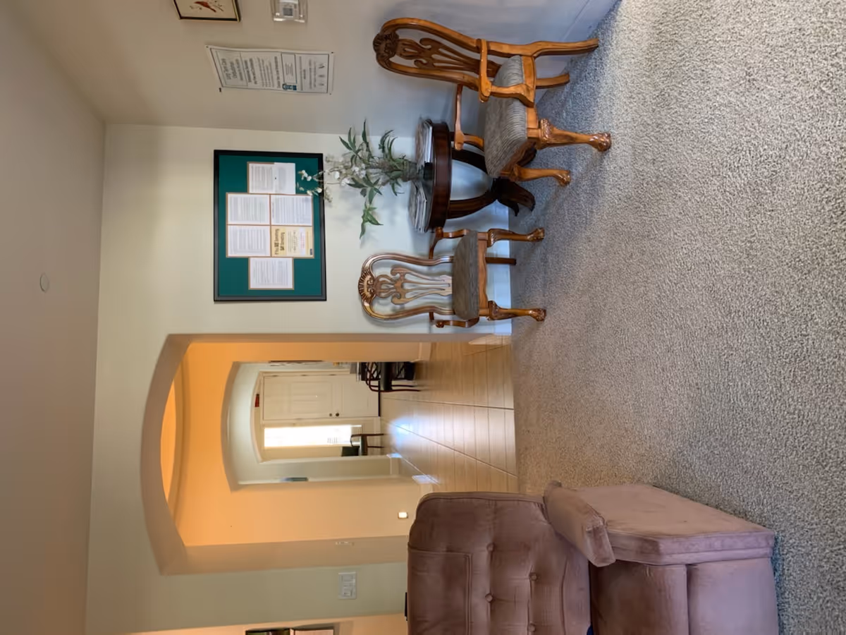 A cozy interior room with beige carpet and cream-colored walls. There are two wooden chairs with patterned cushions and a small round wooden table with a plant on it. A mauve upholstered armchair with a matching ottoman is in the foreground. The room has an arched doorway leading to a tiled hallway with white cabinets and a window letting in natural light. Various framed notices and a thermostat are mounted on the wall.