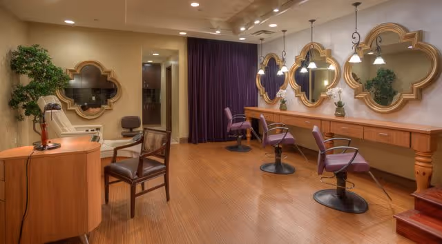 Interior view of a salon area with wooden flooring, three styling chairs in front of a long counter with three decorative mirrors on the wall. There is a small desk with a chair and a hairdryer on the left side, and a purple curtain in the background.