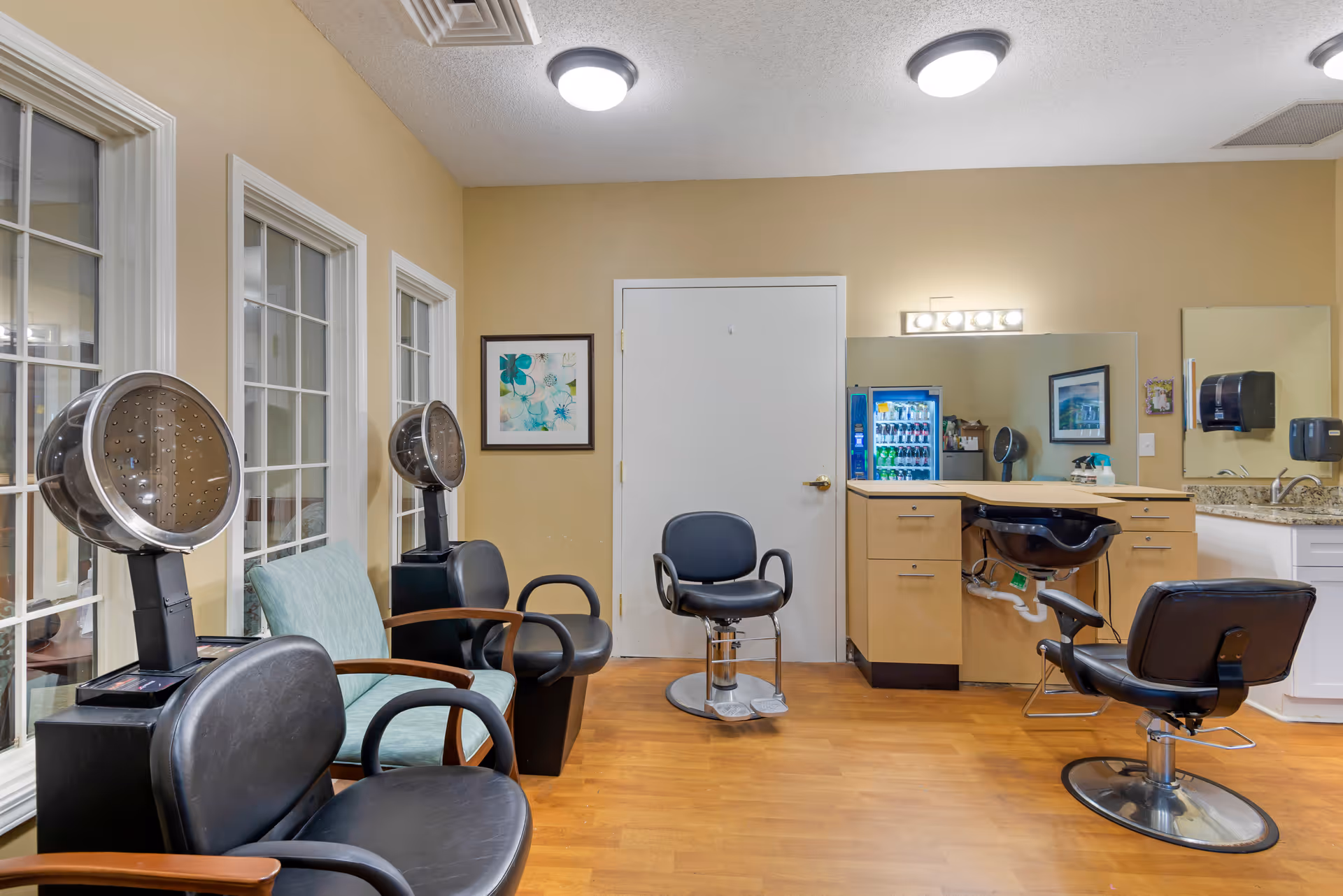 Interior of a hair salon area in a senior living facility with multiple black salon chairs, two hair dryers, a sink for washing hair, a small refrigerator stocked with drinks, and beige walls with framed artwork.
