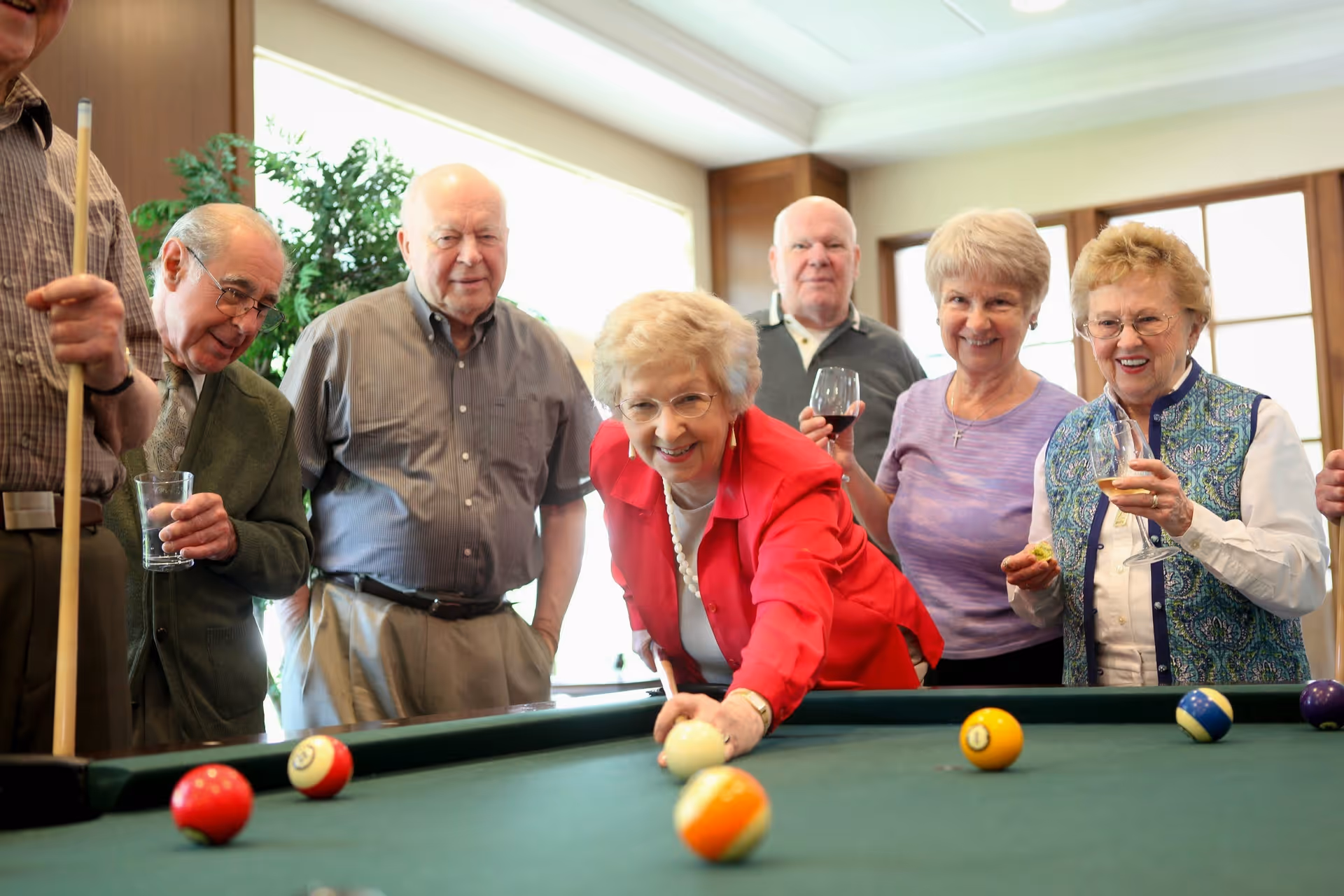 A group of elderly people gathered around a pool table indoors. One woman in a red jacket is leaning forward to take a shot with a pool cue, while others watch and smile. Some are holding drinks. The room is well-lit with large windows and wooden paneling.