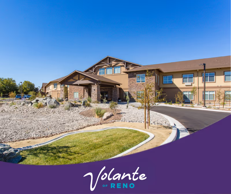 Front exterior of a two-story senior living building with a covered entrance, driveway and rocky landscaping under a clear blue sky, with a purple banner reading 'Volante of Reno' at the bottom.