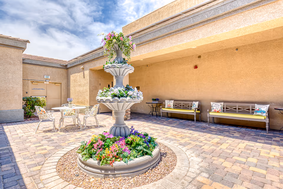 Outdoor courtyard area with a three-tiered stone fountain decorated with colorful flowers in the center. Surrounding the fountain are paved stone walkways, two benches with yellow cushions and decorative pillows, and a white metal table with matching chairs. The walls are beige stucco, and the sky is partly cloudy.