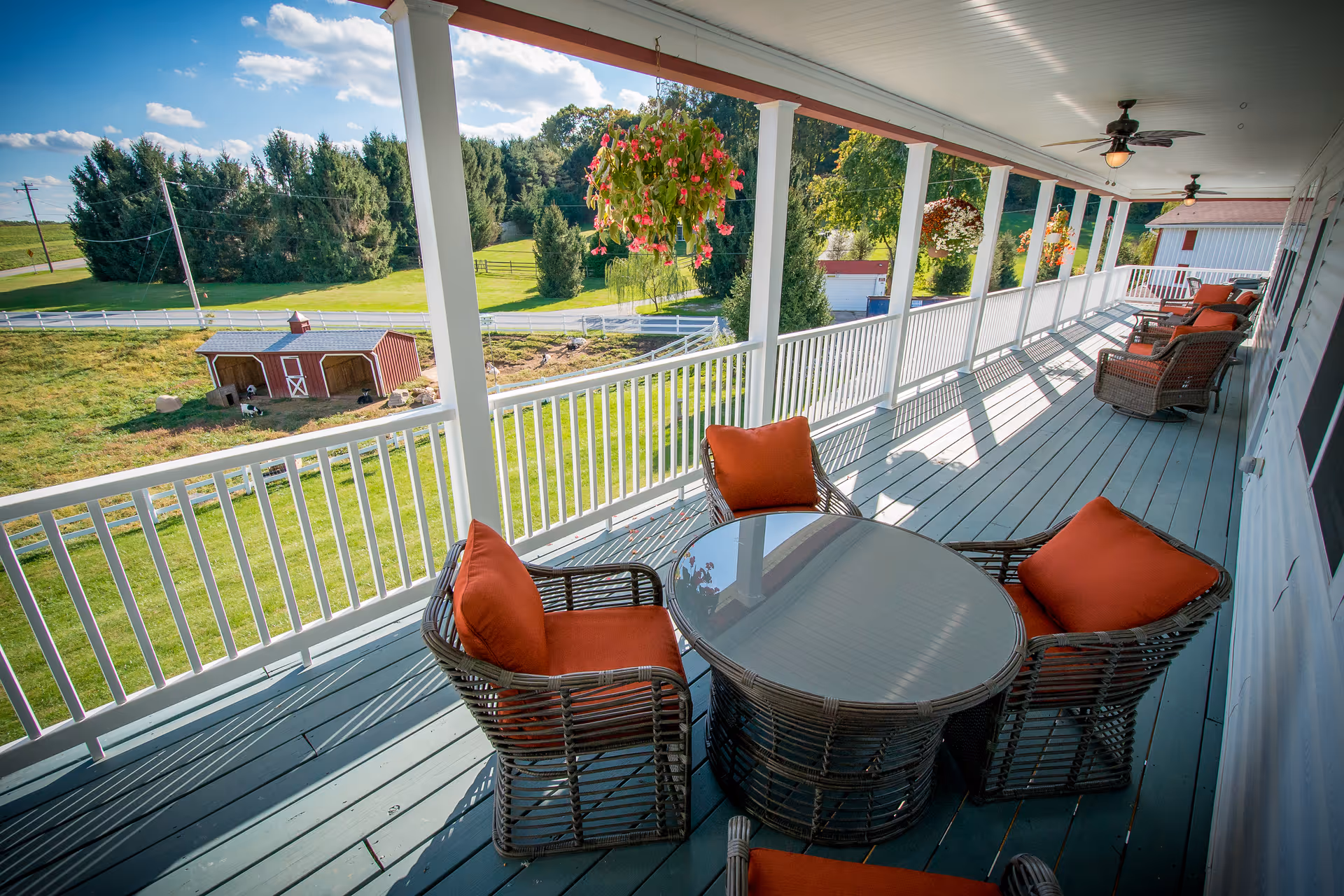 Covered wraparound porch with wicker chairs and a glass-top table overlooking a grassy yard and a small red barn.