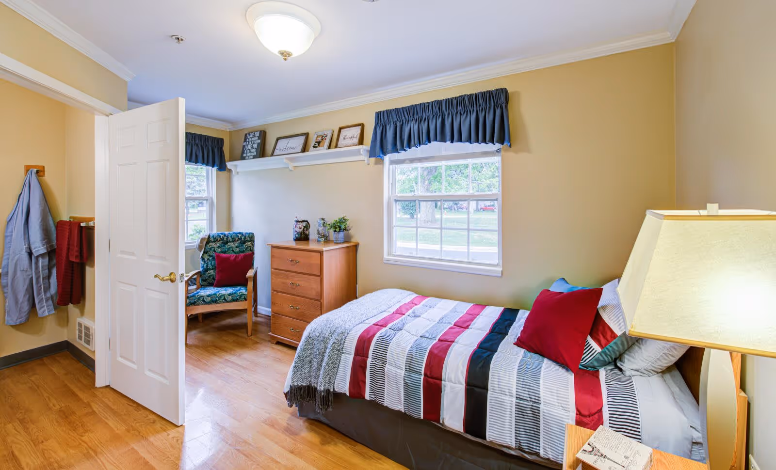 A cozy bedroom with a single bed covered in a striped red, black, white, and gray comforter, and several pillows including a red one. Next to the bed is a wooden nightstand with a lamp and a small box. The room has light yellow walls, two windows with blue valances, a wooden dresser, and a floral upholstered chair with a red pillow. A white door is open, revealing a coat and towel hanging on hooks on the wall.