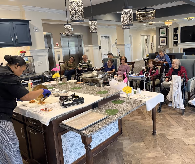 A woman wearing gloves is preparing food on a kitchen island in a senior living facility. Several elderly residents are seated in chairs and wheelchairs in the background, watching the food preparation. The room has pendant lights hanging from the ceiling, a granite countertop, and a cozy seating area with framed pictures on the wall.