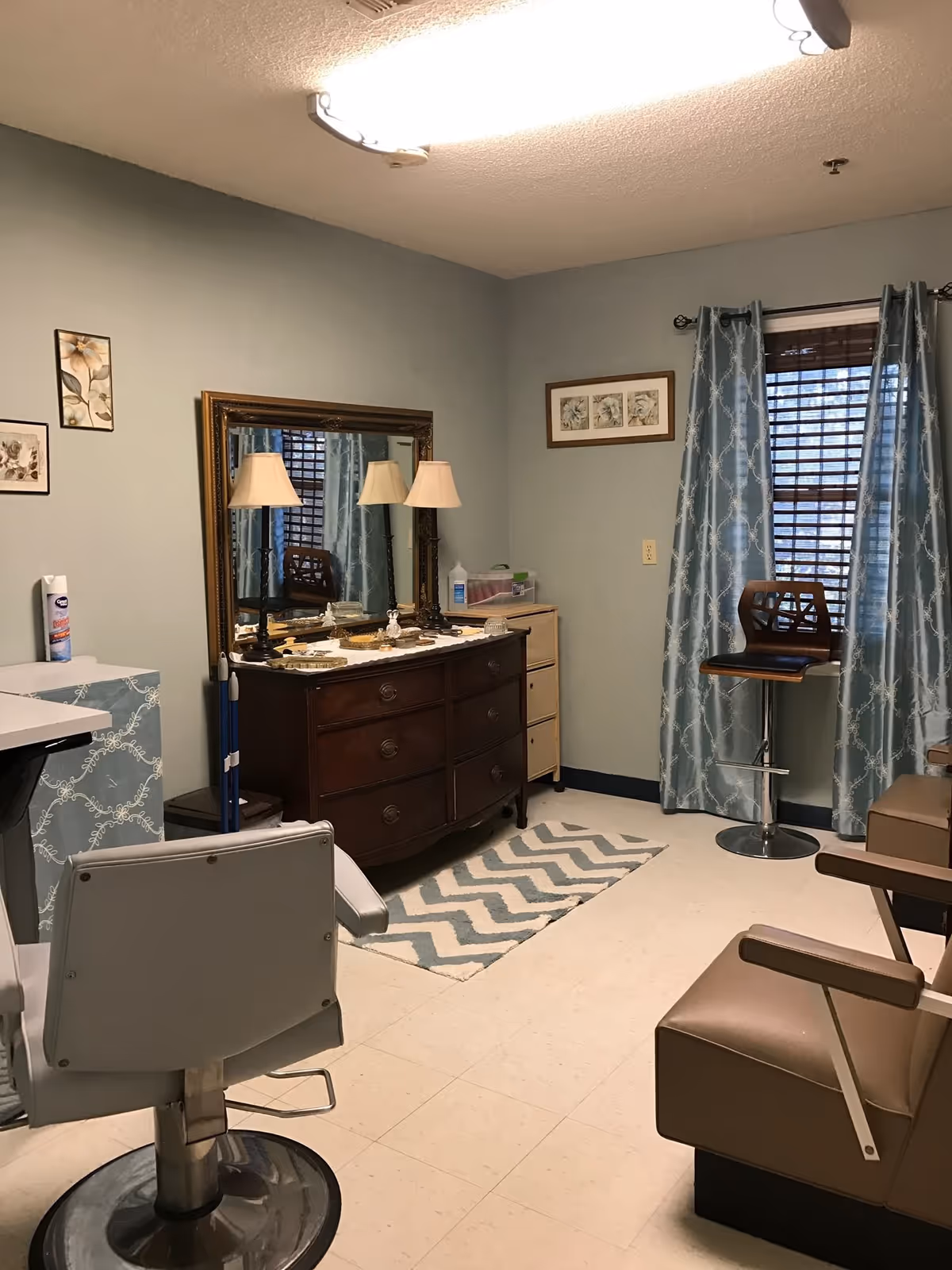 Interior room with light blue walls and tiled floor, featuring a large wooden dresser with a mirror and two lamps on top, a chevron patterned rug, a tall chair near a window with blue patterned curtains, and two beige armchairs. Various framed pictures hang on the walls.
