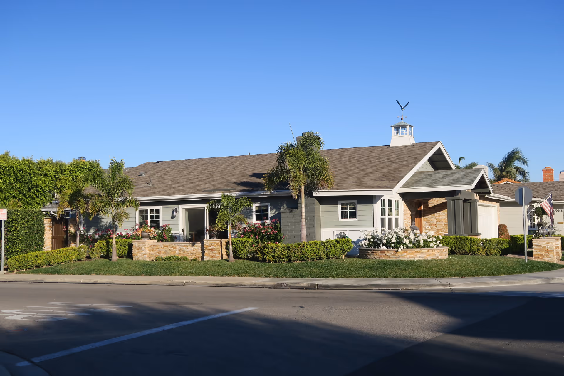 Exterior view of a single-story senior care facility building with a well-maintained garden featuring palm trees, bushes, and flowers. The building has a gray roof, light-colored walls, and a small cupola with a weather vane on top. The sky is clear and blue.