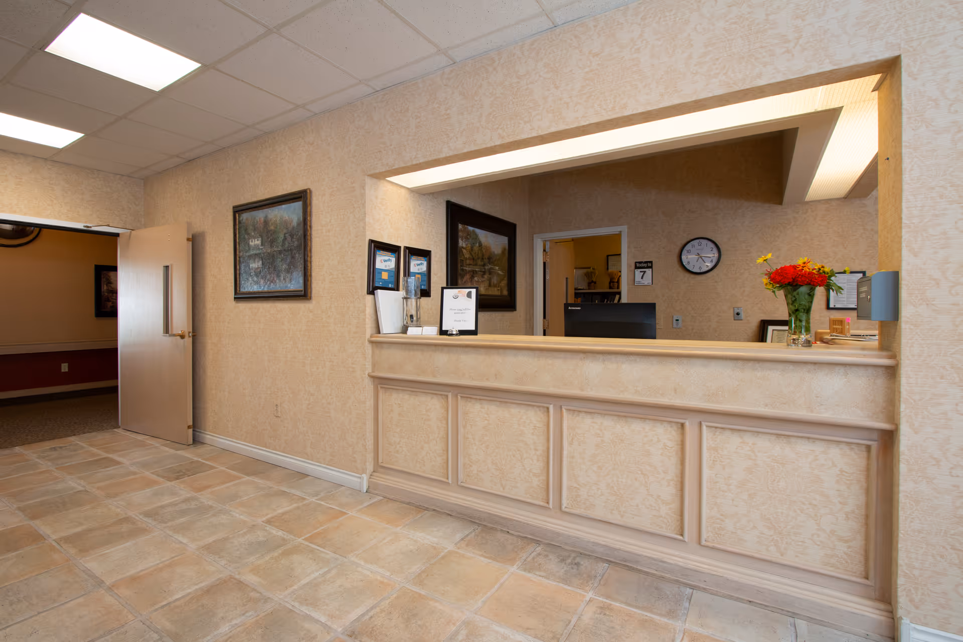 Reception area with a beige tiled floor and beige patterned wallpaper. There is a wooden reception desk with a computer monitor, a vase with red and yellow flowers, and framed pictures on the walls. A door is open to the left, leading to another room with darker walls and carpeted floor.