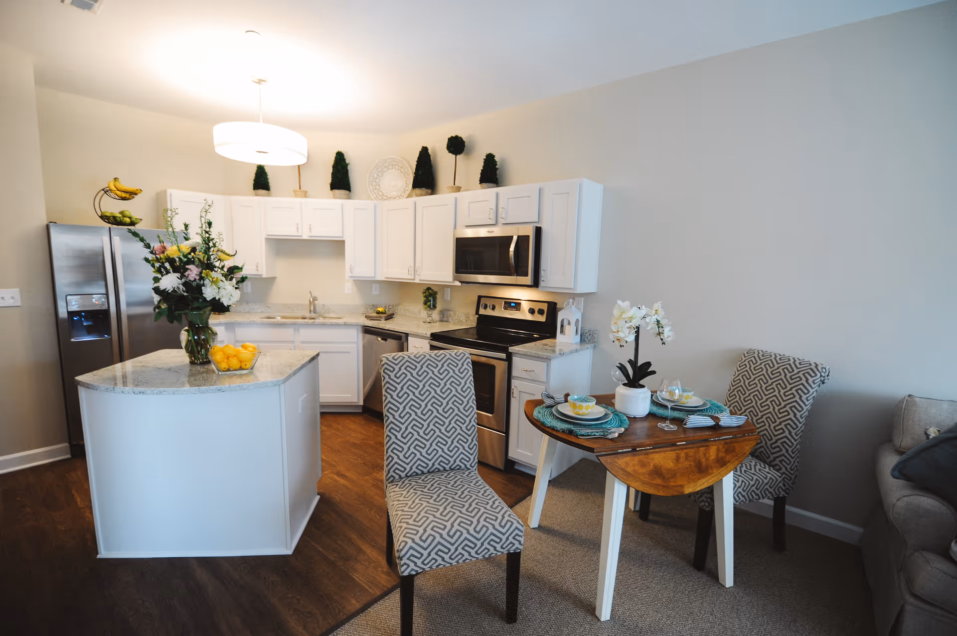 A modern kitchen and dining area featuring white cabinets, stainless steel appliances including a refrigerator, microwave, and stove. There is a kitchen island with a vase of flowers and a bowl of lemons. The dining area has a small wooden table set for two with patterned chairs and a white orchid centerpiece. The floor is a combination of wood and carpet.