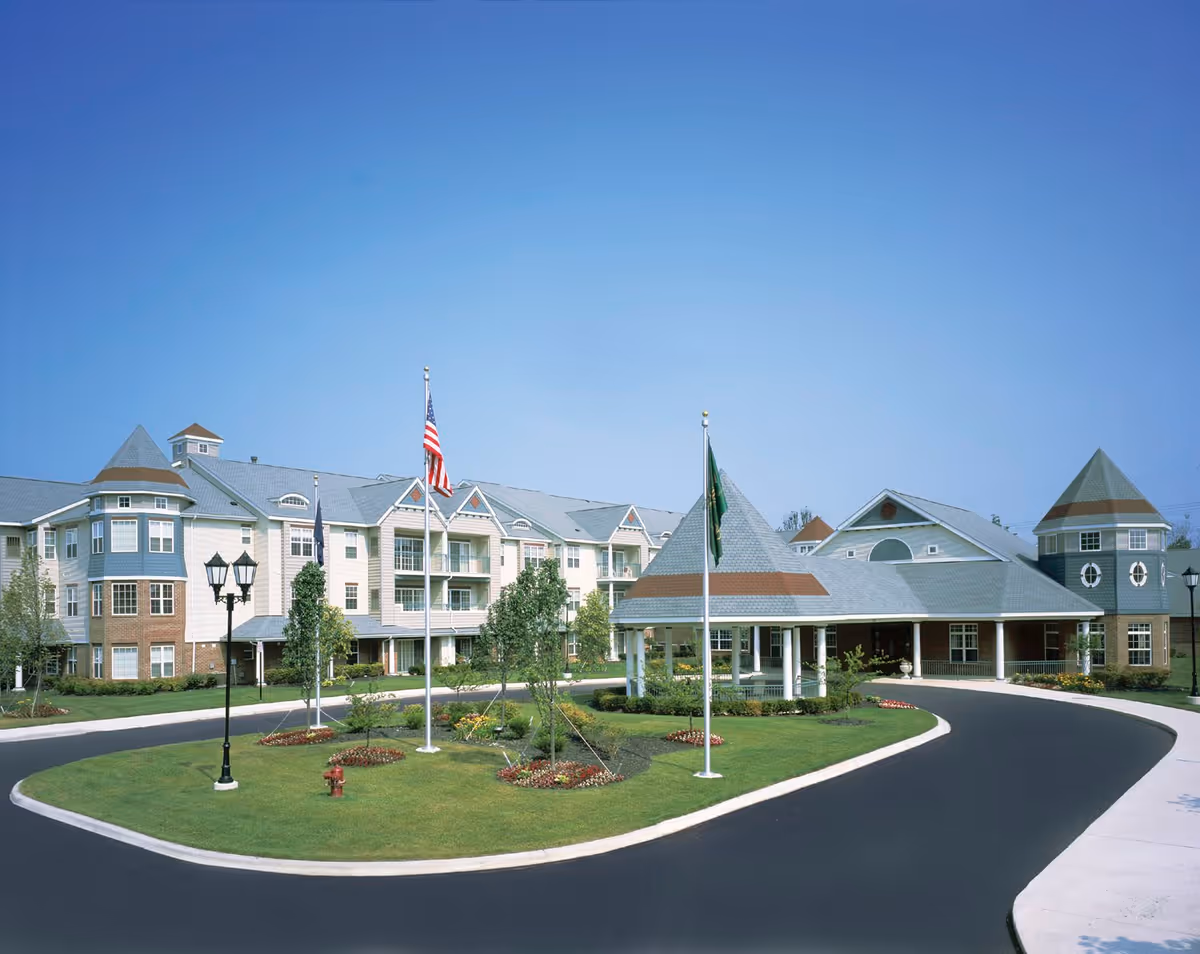 Exterior view of Waltonwood Cherry Hill senior living facility with a circular driveway, landscaped lawn, flagpoles with American and green flags, and a multi-story building featuring turrets and balconies under a clear blue sky.