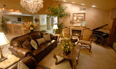 A cozy living room area with a brown leather sofa and matching armchair, a marble coffee table with a floral arrangement, two upholstered chairs near a fireplace, a grand piano in the corner, a large chandelier hanging from the ceiling, and decorative plants and lamps providing warm lighting.