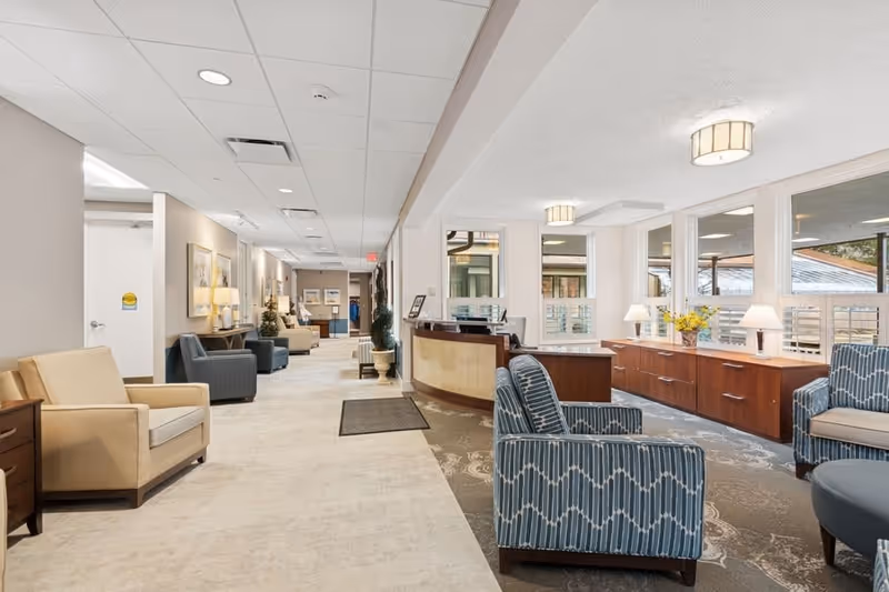 A bright and spacious senior living facility interior with a long hallway lined with beige and blue armchairs, side tables, and lamps. On the right side, there is a reception desk with large windows behind it, letting in natural light. The area is decorated with framed artwork, plants, and flowers, creating a welcoming atmosphere.
