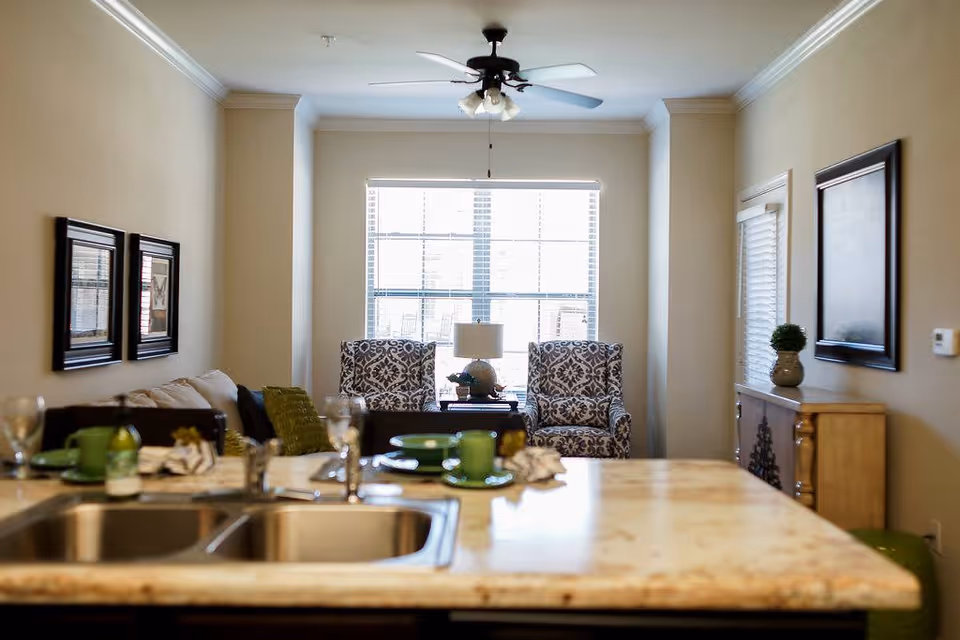 View from a kitchen island and sink into a bright living room with two patterned armchairs by a large window, a ceiling fan, and a sofa with framed art on the walls.