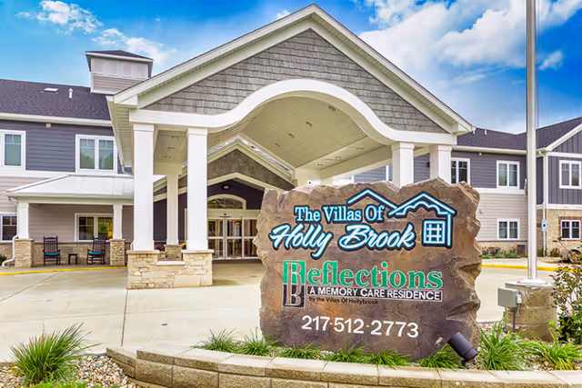 Covered entrance and porte-cochere of The Villas of Holly Brook with a large stone sign for "Reflections A Memory Care Residence" in the foreground.