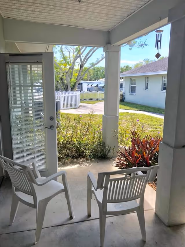 View from a covered patio area with two white plastic chairs facing outward toward a garden with green grass, bushes, and a white picket fence. A white door with glass panes and blinds is open to the patio. In the background, there is a white building and some trees under a clear blue sky.