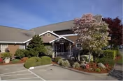 Single-story brick senior living building entrance with a covered porch, landscaped shrubs and trees, and a small parking area under a blue sky.
