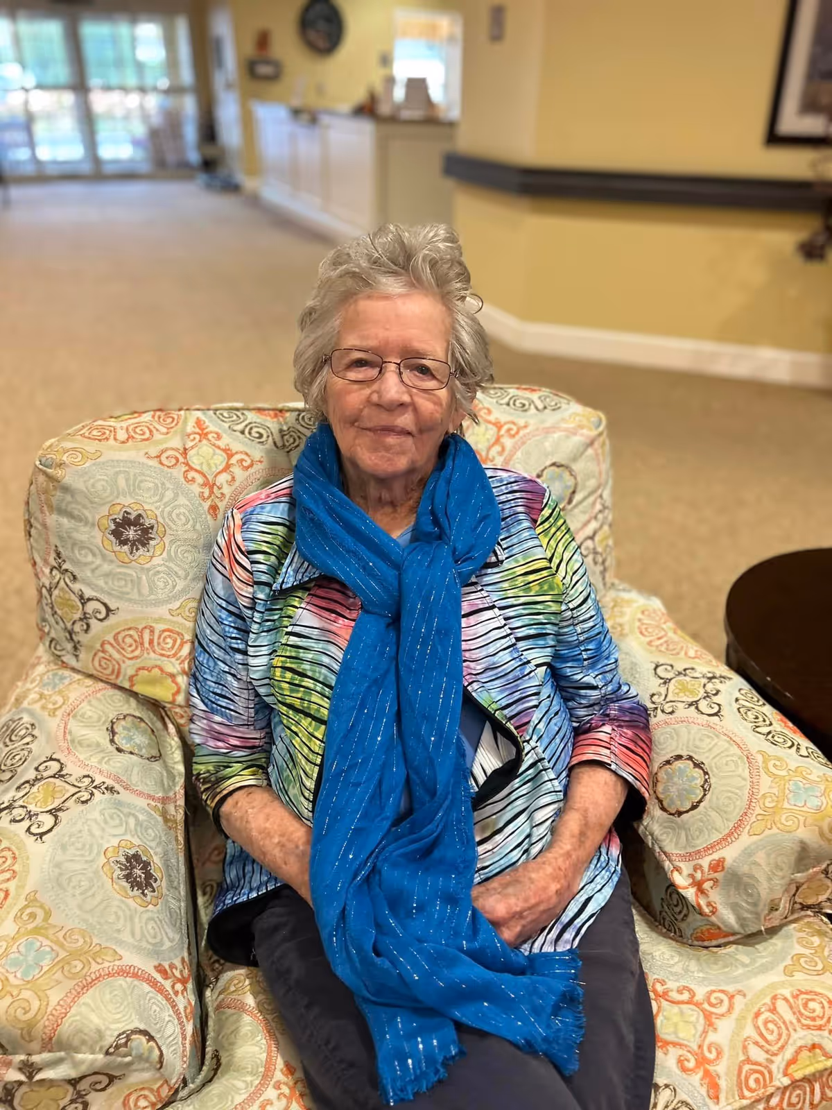 An elderly woman with short gray hair and glasses is sitting on a patterned armchair. She is wearing a colorful striped shirt and a bright blue scarf. The background shows a spacious, well-lit interior with beige walls and carpet, and a reception desk in the distance.