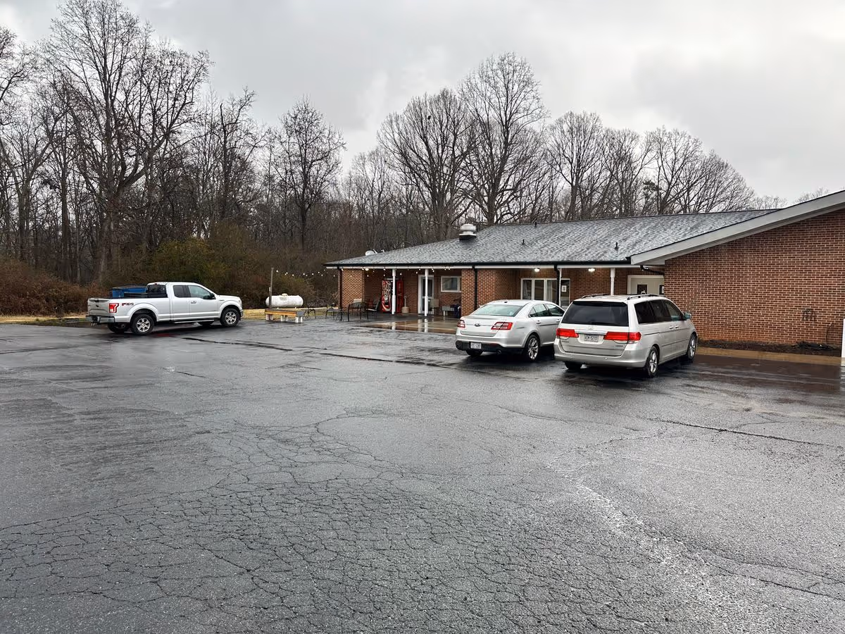 Brick single-story assisted living building with a wet parking lot and several parked vehicles in front under an overcast sky.