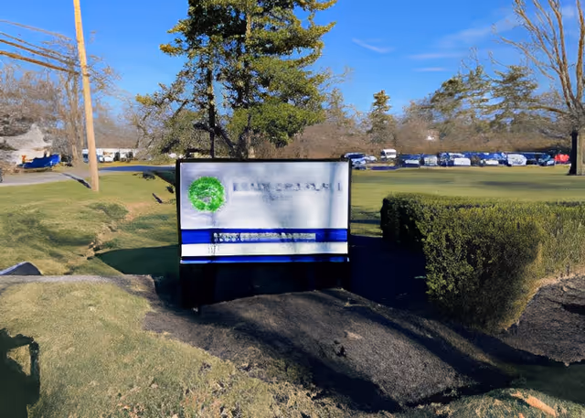 Outdoor view of a grassy area with a sign for Bradford Place in the center, surrounded by trees, bushes, and a parking lot with cars in the background under a clear blue sky.