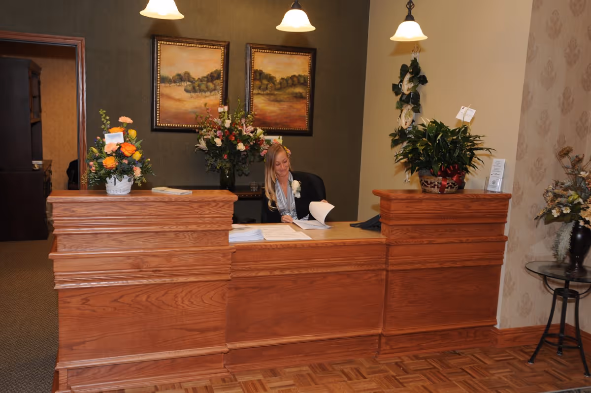 A receptionist sits behind a wooden front desk in a decorated lobby with floral arrangements and framed paintings.