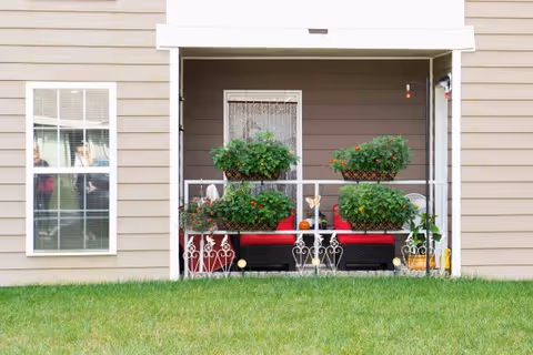 Covered ground-level patio with red cushioned seating and multiple potted plants in front of a beige building facade and lawn.