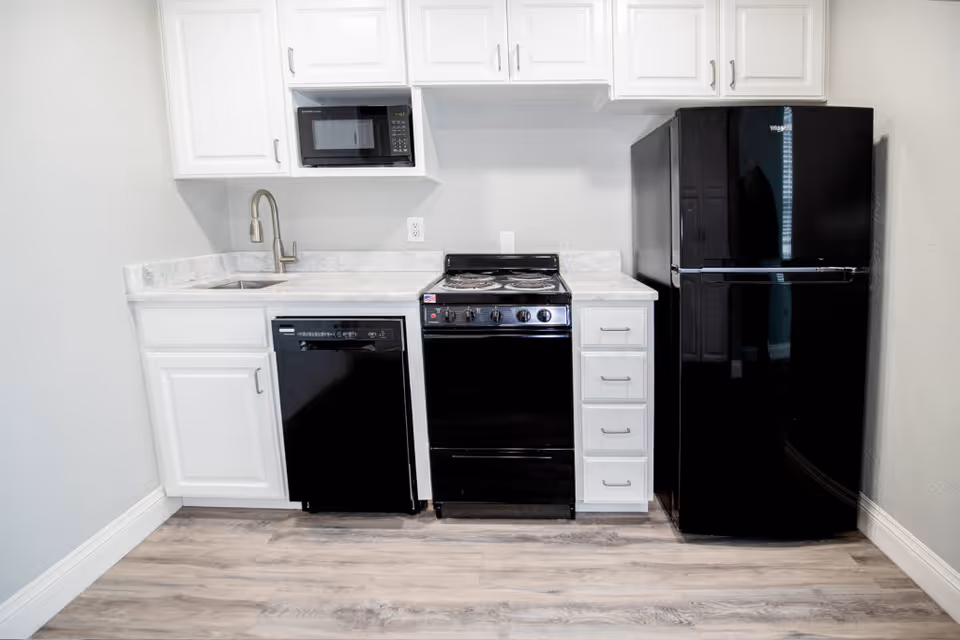 A small kitchen area with white cabinets and marble countertops. The kitchen includes a black microwave, black dishwasher, black stove with four burners, and a black refrigerator. The floor is light wood, and the walls are painted white.