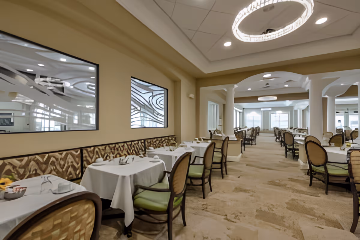 A spacious dining room with multiple tables covered in white tablecloths, each set with plates, glasses, and napkins. The room features cushioned chairs with wooden frames and green seats, decorative wall mirrors, beige walls, and a carpeted floor. Large windows at the far end allow natural light to fill the space, and modern circular light fixtures hang from the ceiling.