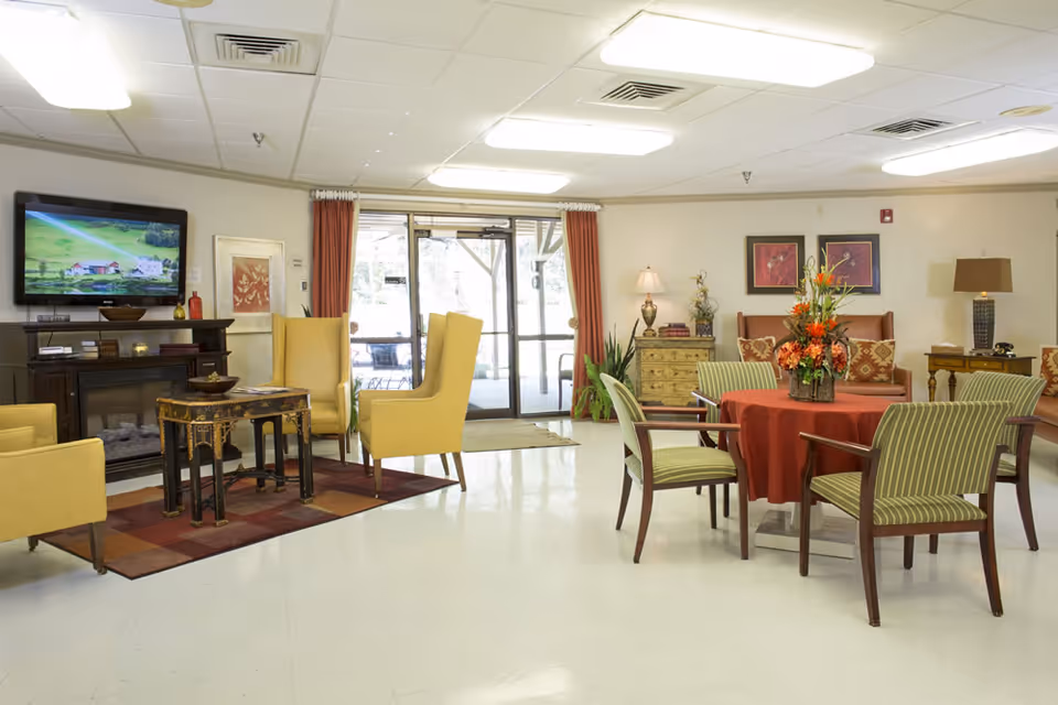 A bright and spacious common area in a senior living facility with yellow armchairs arranged around a small table on a patterned rug, a wall-mounted TV above a fireplace, a round table covered with a red tablecloth and decorated with a floral centerpiece, green striped chairs around the table, and additional seating and lamps along the walls. Large glass doors with red curtains lead outside.
