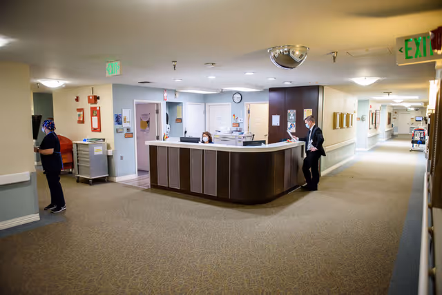 A reception desk area inside a senior living facility with two people wearing masks, one standing behind the desk and another leaning on it. The hallway extends to the right with framed pictures on the wall and medical equipment visible in the distance. The area is well-lit with ceiling lights and has beige walls and carpeted floors.