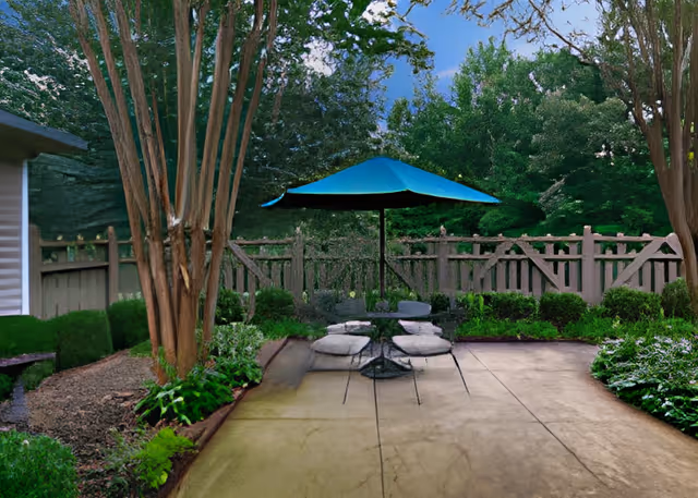 Outdoor patio area with a round table and four chairs, each with a cushion, under a large blue umbrella. The patio is surrounded by green bushes, trees, and a wooden fence in the background.
