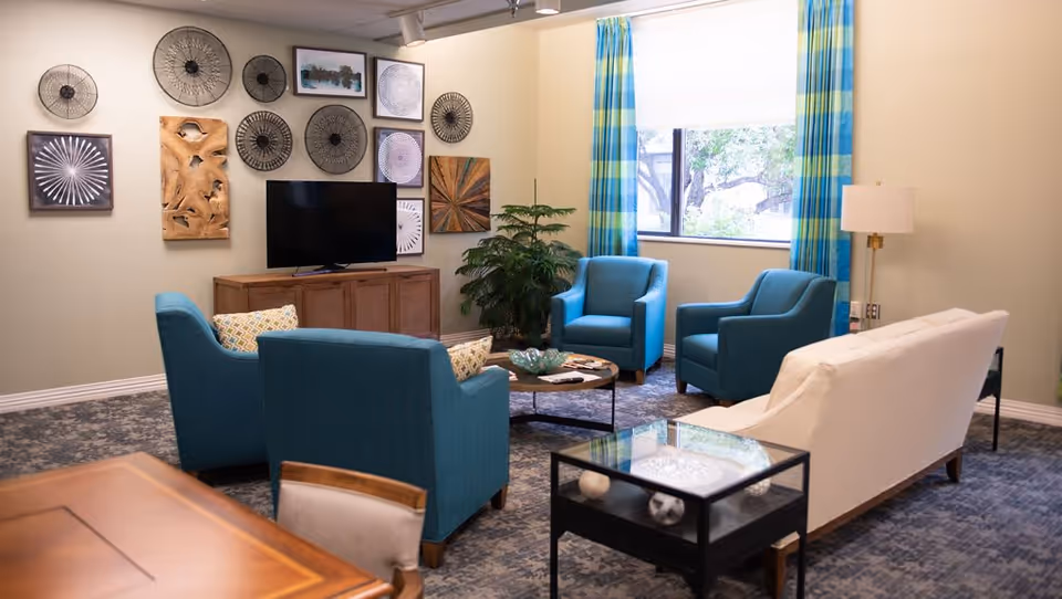 A cozy common living room with teal armchairs and a white sofa arranged around a coffee table, a TV on a cabinet with decorative wall art, and a window with blue plaid curtains.
