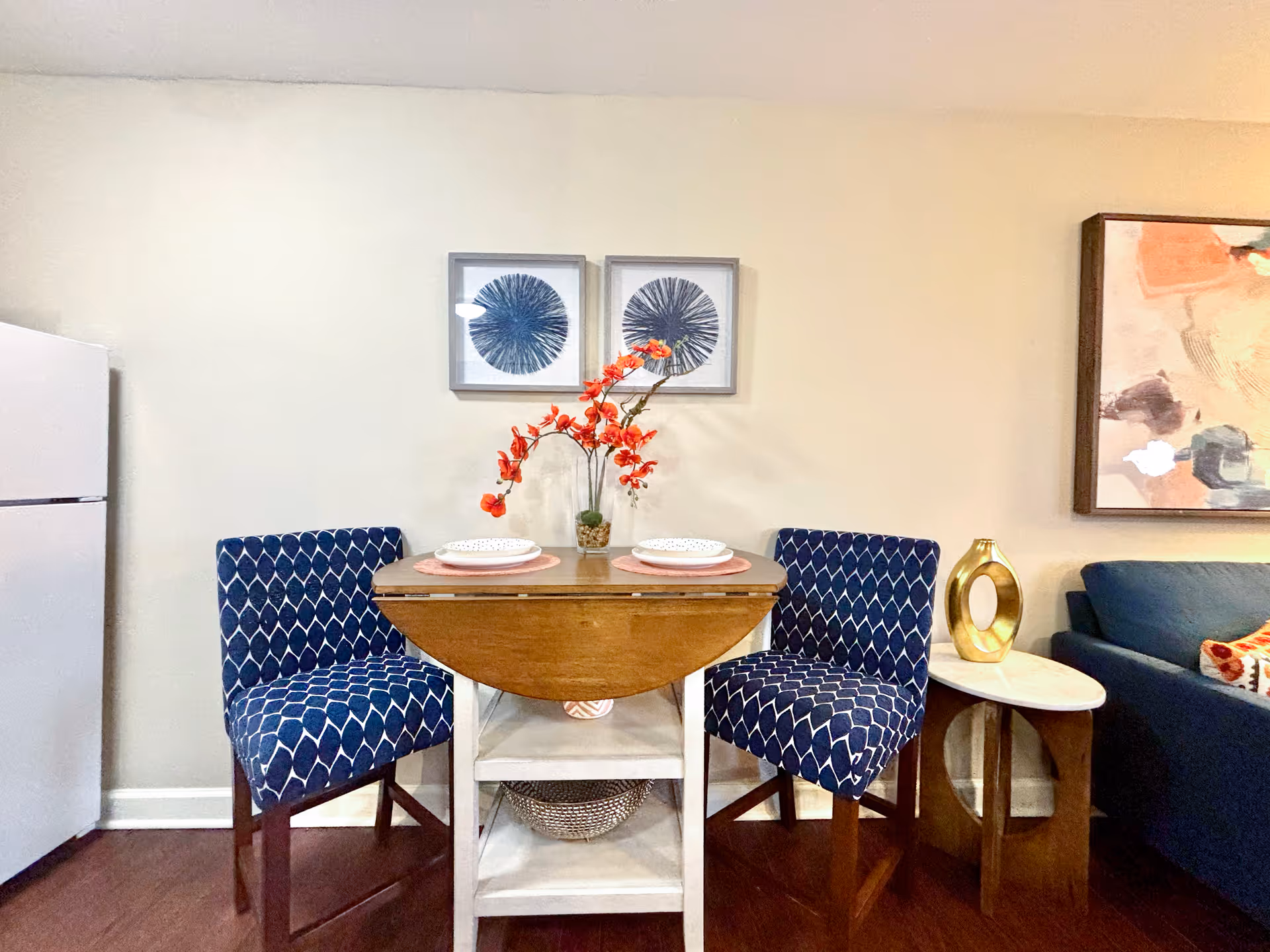 Small dining area with a wooden drop-leaf table set for two with white plates on pink placemats, flanked by two blue patterned chairs. Behind the table are two framed abstract blue artworks on a beige wall. To the left is a white refrigerator, and to the right is a blue couch with a side table holding a gold decorative object and a colorful abstract painting above.