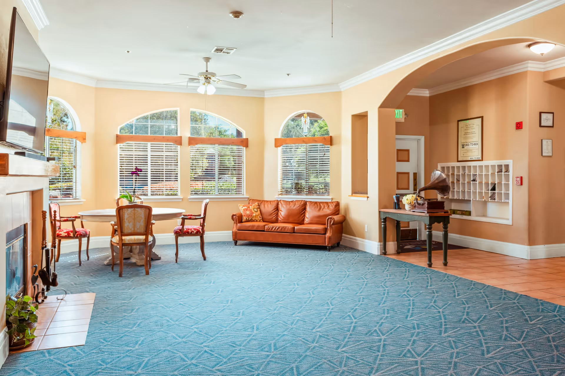 A bright and spacious assisted living common area with large arched windows letting in natural light. The room features a brown leather sofa, a round wooden table with three upholstered chairs, a fireplace with a mounted TV above it, and a small table with a vintage gramophone. The walls are painted beige, and the floor is covered with a blue patterned carpet and some terracotta tiles near the entrance.