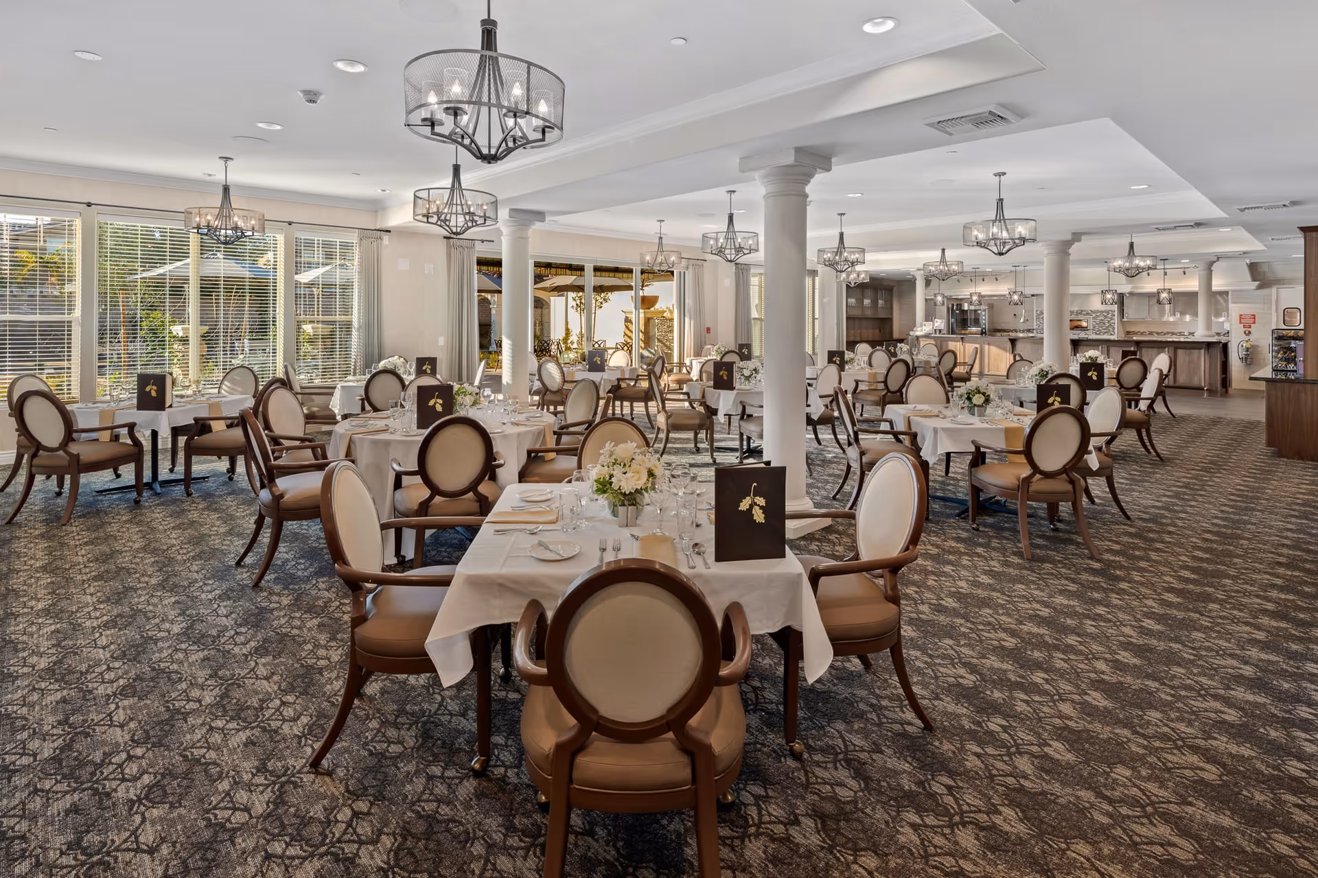 Elegant senior living dining room with round tables set with white linens and cushioned chairs under chandeliers and large windows.