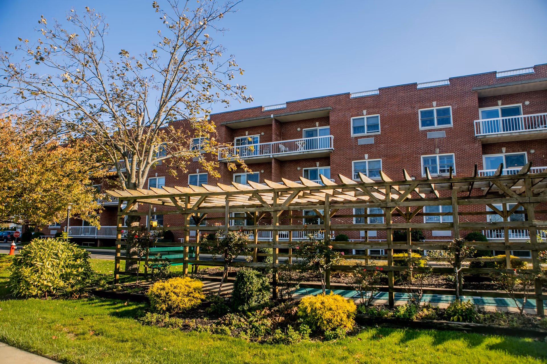 Exterior view of a multi-story brick building with balconies, surrounded by a garden area featuring a wooden pergola, green shrubs, and trees under a clear blue sky.