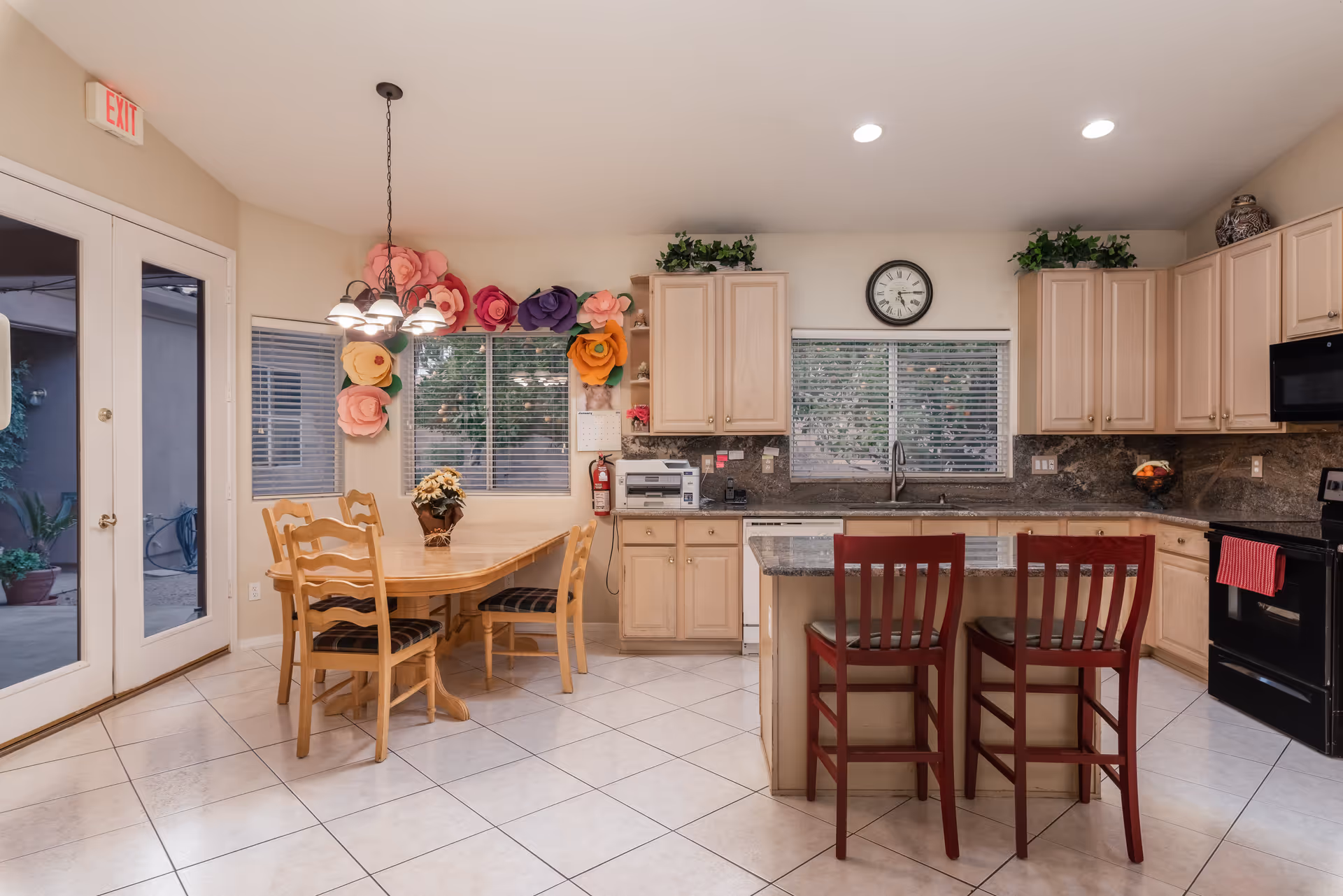A bright kitchen and dining area with light wood cabinets, granite countertops, and tiled floor. The kitchen features a black stove, microwave, dishwasher, and a kitchen island with two red chairs. The dining area has a wooden table with four chairs and a floral centerpiece. Large windows with blinds and colorful paper flower decorations are on the wall behind the dining table. A clock is mounted above the kitchen sink, and an exit door with glass panels is visible on the left side.