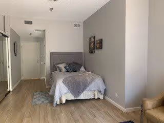 A small bedroom with a single bed against a gray wall, decorated with two framed pictures above the bed. The bed has gray and white bedding with multiple pillows. The room has light wood flooring, a small rug near the bed, and a beige armchair partially visible on the right side. A hallway and a door are visible in the background.