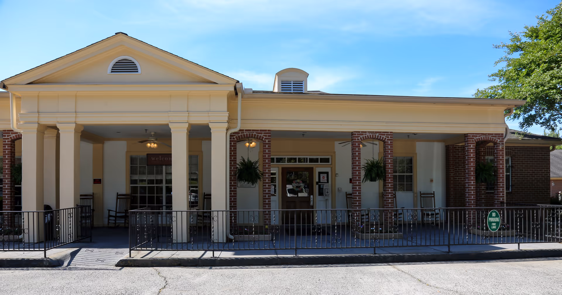 Front exterior view of Green Acres Health & Rehabilitation building with a covered porch supported by columns, rocking chairs, hanging plants, and a clear blue sky above.