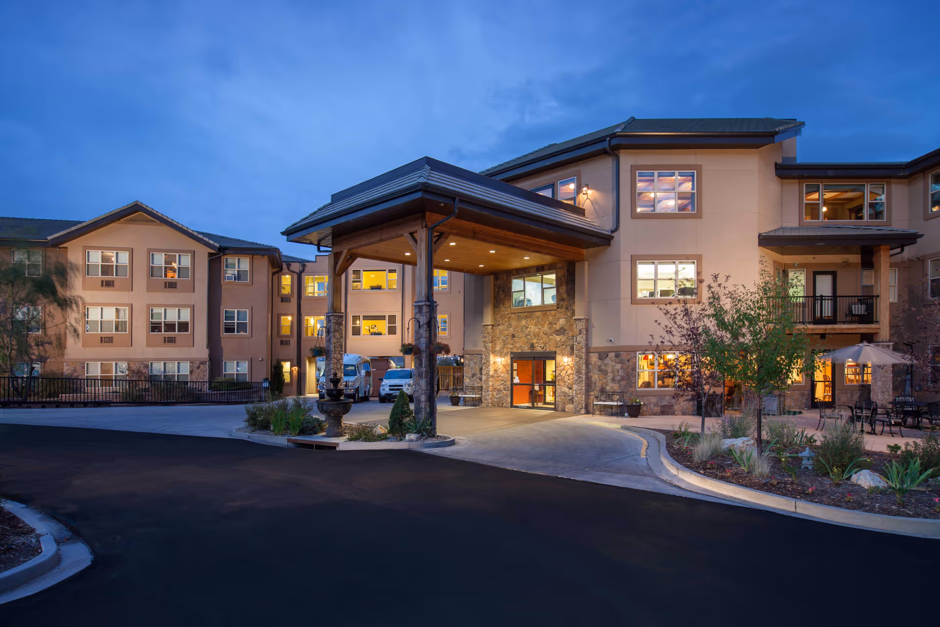 Exterior view of The Palisades at Broadmoor Park senior living facility at dusk, showing a multi-story building with stone and beige walls, a covered entrance with wooden beams, lit windows, landscaped garden areas, and a driveway.