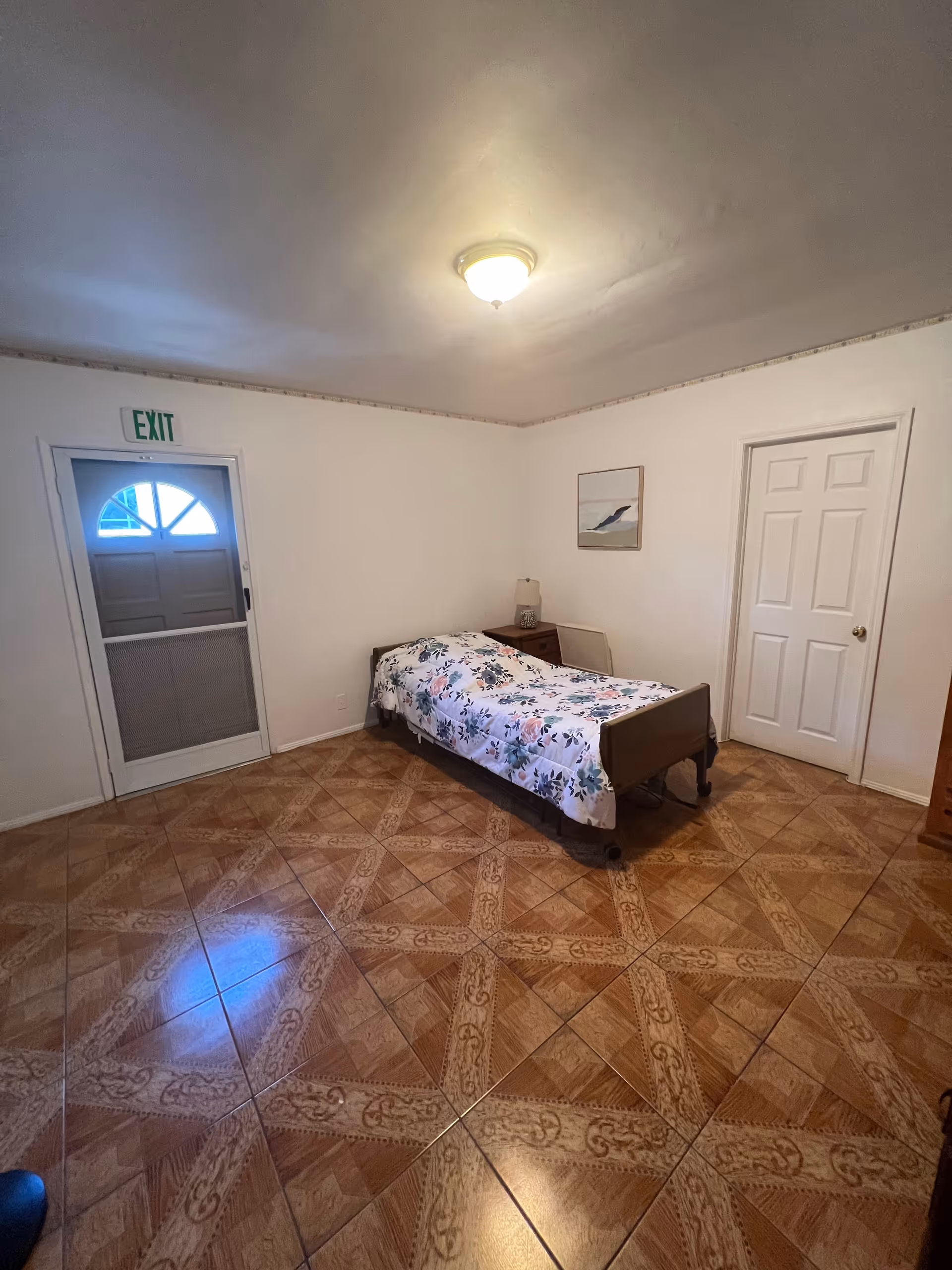 A simple bedroom with a single bed covered in a floral bedspread, a wooden nightstand with a lamp, a framed picture on the wall, a closed white door, and an exit door with a window. The floor has patterned brown tiles and the ceiling has a single light fixture.