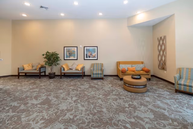 Spacious furnished common room with sofas, chairs, patterned carpet, framed artwork, and recessed ceiling lights.