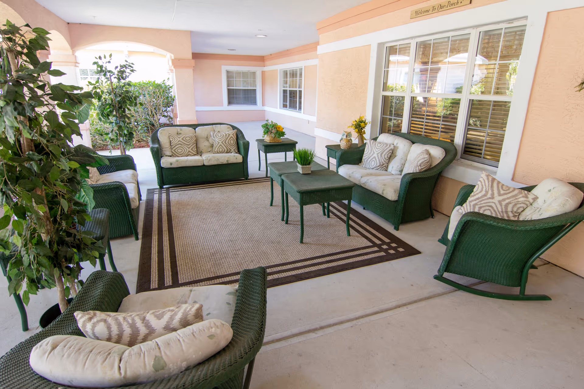 Covered outdoor porch area with green wicker furniture including cushioned chairs, loveseats, and tables arranged on a large beige and brown rug. There are decorative plants and flowers on the tables, and windows with white frames on the peach-colored wall behind the seating area.