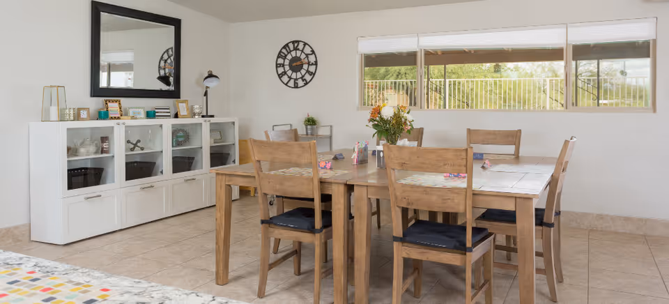 Bright dining room with a wooden table and six chairs, a white sideboard beneath a mirror, wall clock, and a long window.