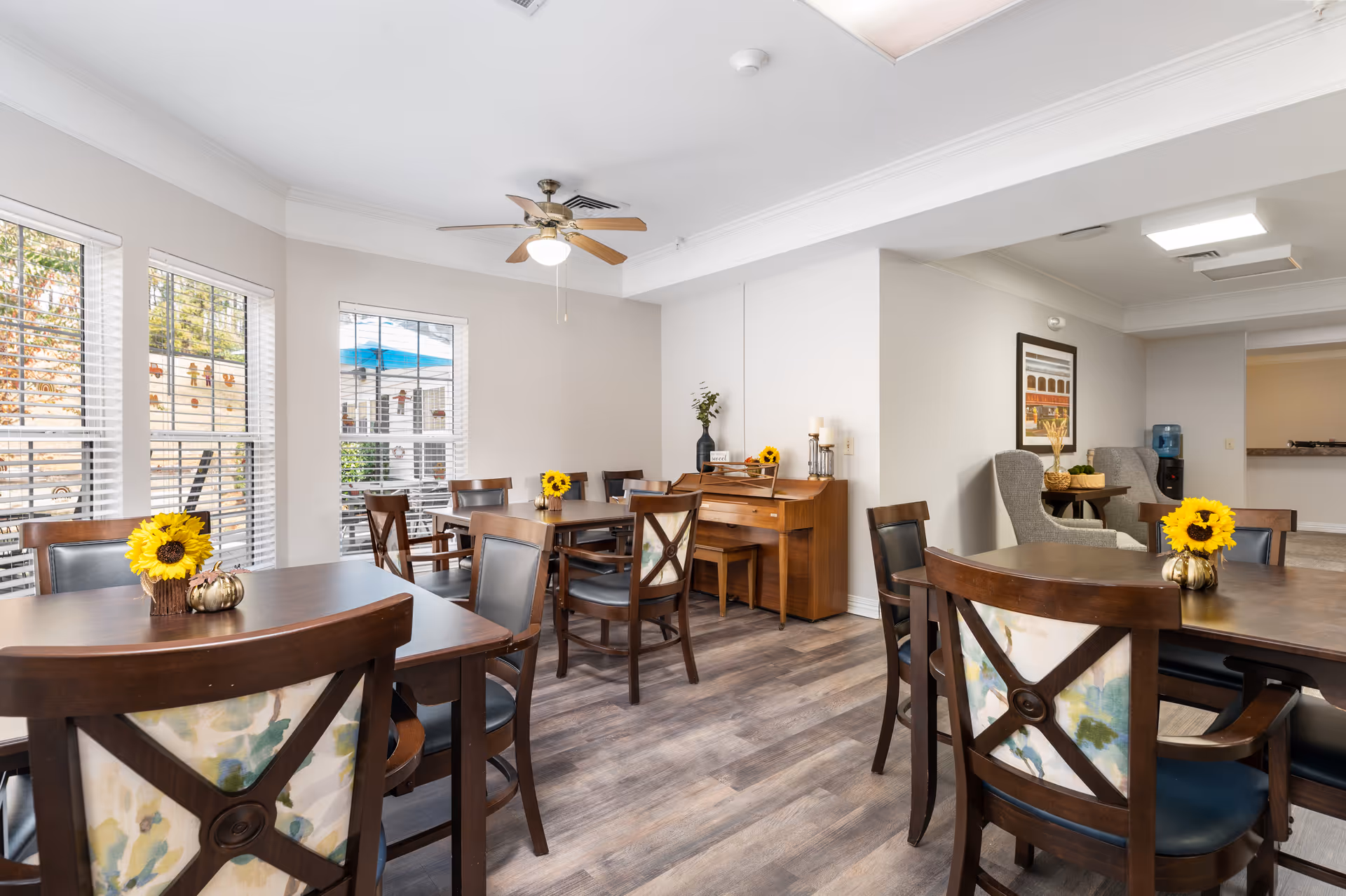 Bright communal dining room with wooden tables and chairs, sunflower centerpieces, large windows, a ceiling fan, and a piano against the wall.