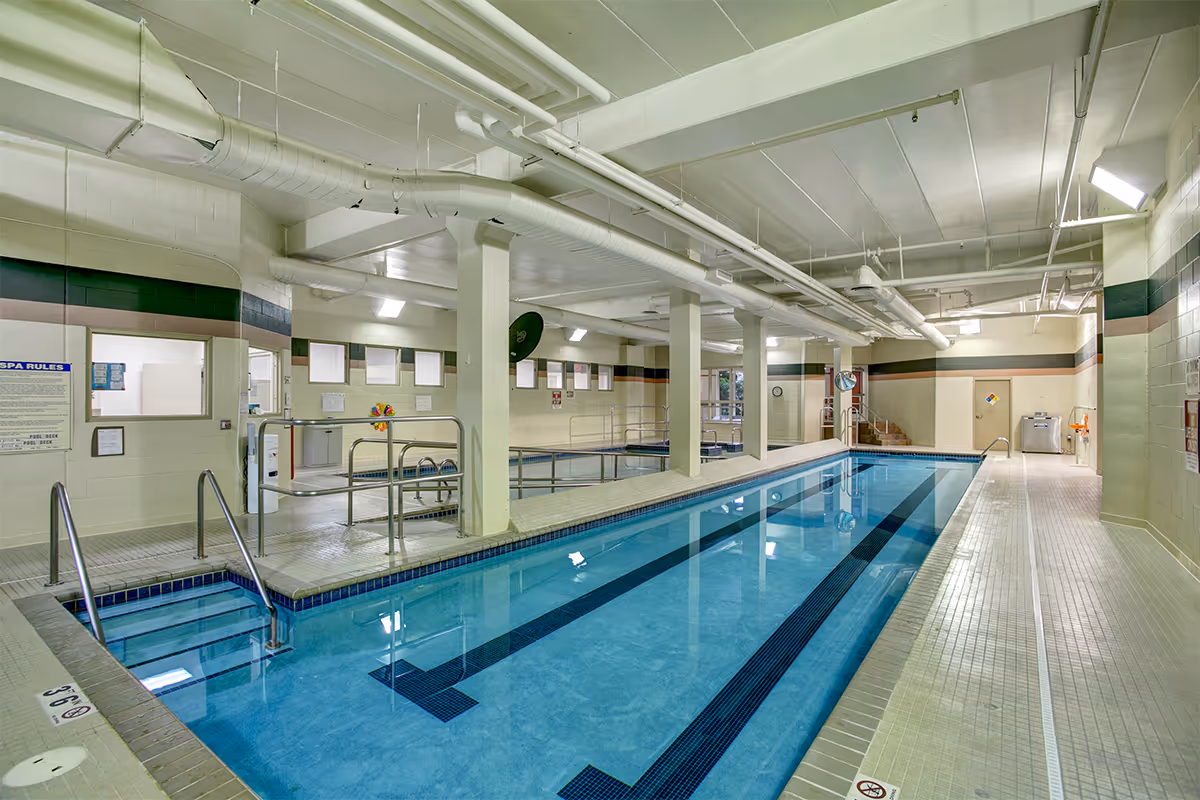 Indoor swimming pool area with clear blue water, handrails, tiled floor, and white ceiling with exposed pipes. The pool is long and narrow, with a smaller pool or hot tub adjacent to it. Walls are painted in light colors with a dark stripe near the ceiling. There are windows and safety signs on the walls.