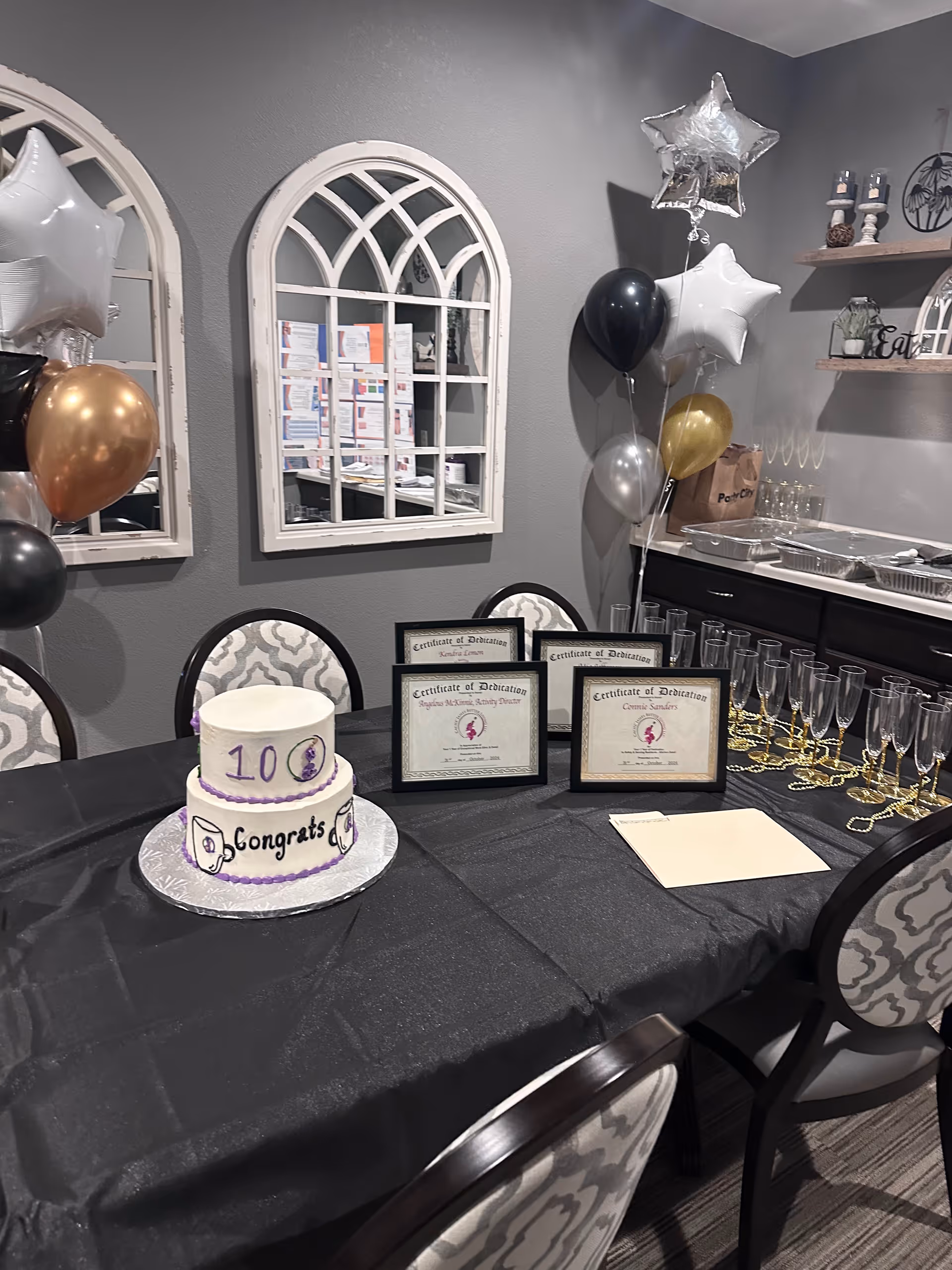A decorated table in a room with a black tablecloth, a two-tiered white cake with purple trim and the word 'Congrats' written on it, four framed certificates of dedication, several empty champagne glasses, and gold bead necklaces. Behind the table are gray walls with two arched mirrors, a cluster of black, gold, silver, and white star-shaped balloons, and shelves with decorative items.