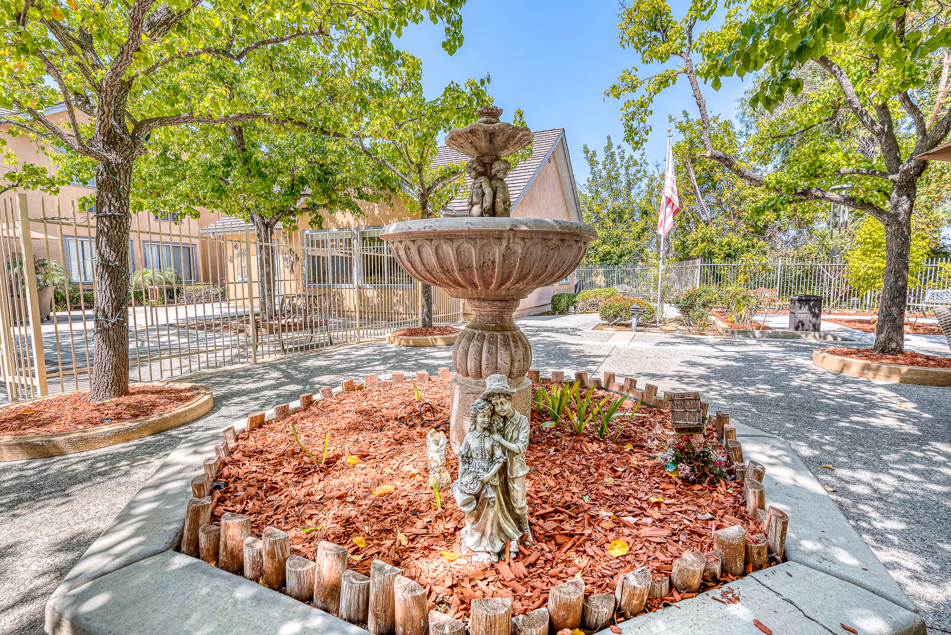 Outdoor courtyard featuring a decorative tiered fountain and statue amid mulched plant beds, trees, and the facility building.