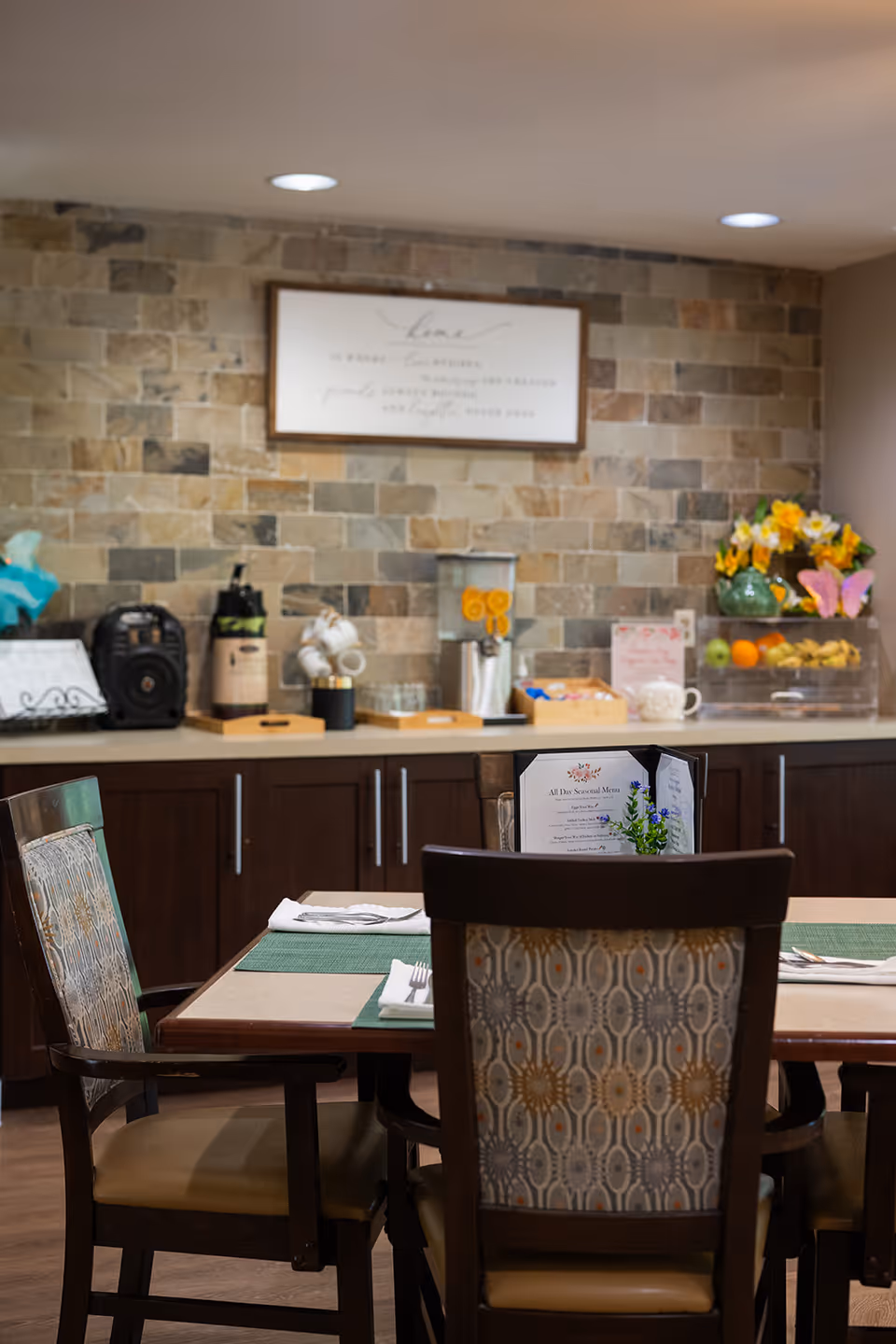 Dining area with a table set for four, featuring patterned cushioned chairs and green placemats. In the background, a countertop with a stone tile backsplash holds a beverage dispenser with orange slices, a coffee station, a fruit tray, and a vase with yellow flowers. A framed sign is mounted on the wall above the countertop.