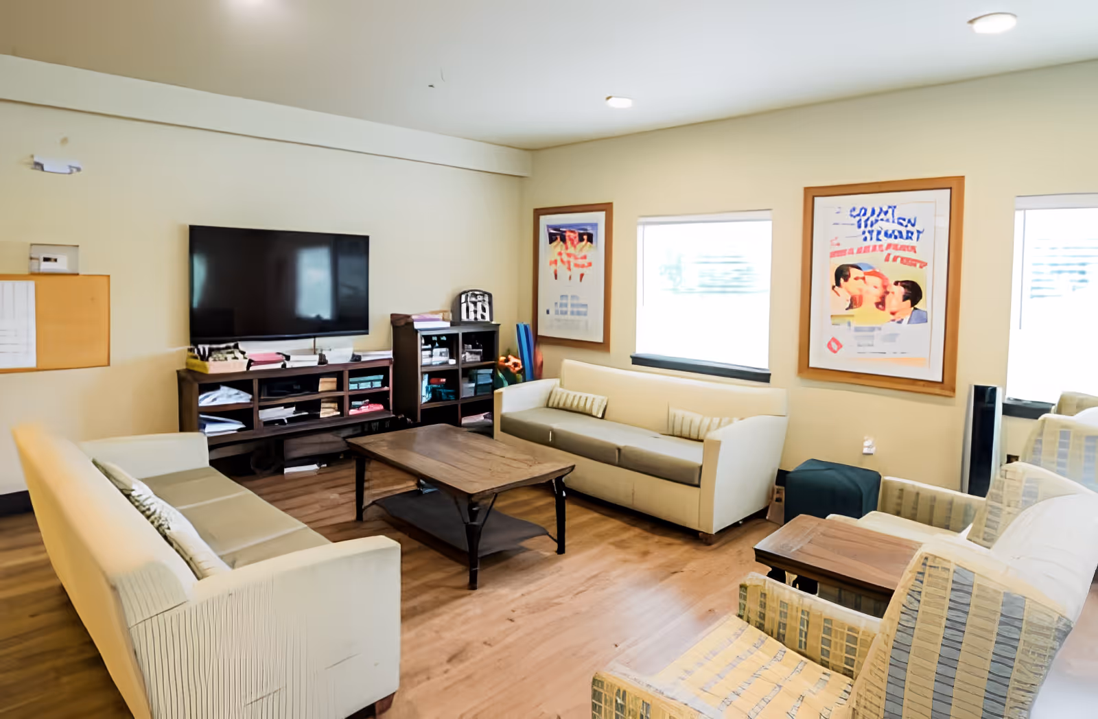 A bright communal lounge with sofas, armchairs, a coffee table, TV and shelving in a senior living facility.